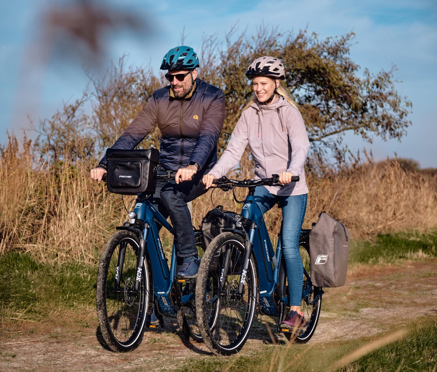 Zwei Personen fahren mit Fahrrädern auf einem Feldweg, wobei das vordere Fahrrad eine Fischer Gepäckträgertasche PLUS Cita trägt.