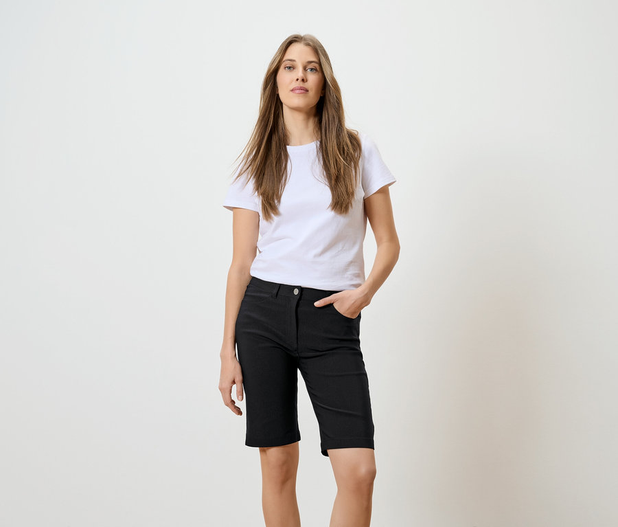 Frau mit langen braunen Haaren, weißem T-Shirt und schwarzen Shorts mit einer Hand in der Tasche vor einem weißen Hintergrund.