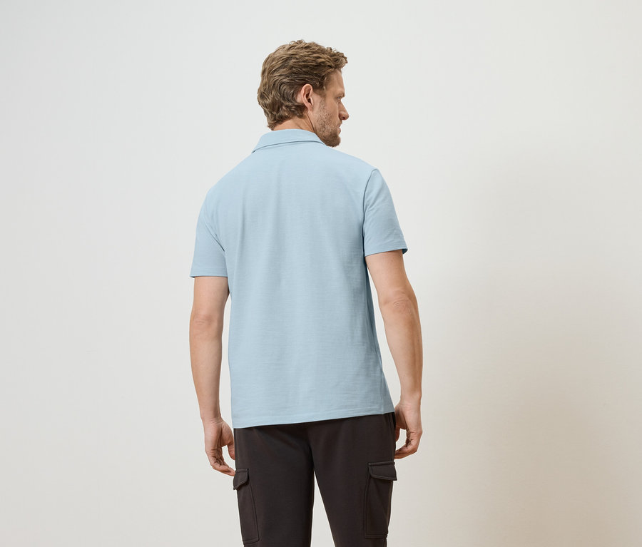 Mann von hinten in hellblauem Poloshirt und dunkler Cargohose vor hellem Hintergrund.