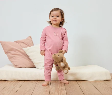 Ein lächelndes Kleinkind im rosa Schlafanzug hält einen Teddybären vor einem Bett mit Kissen.