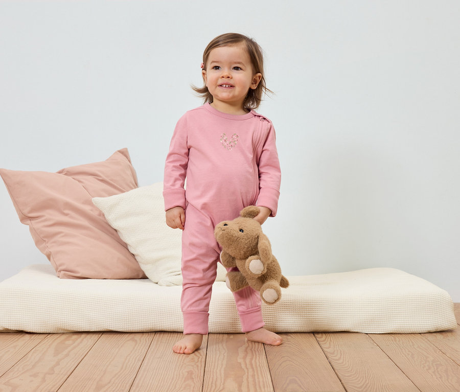 Ein lächelndes Kleinkind im rosa Schlafanzug hält einen Teddybären vor einem Bett mit Kissen.