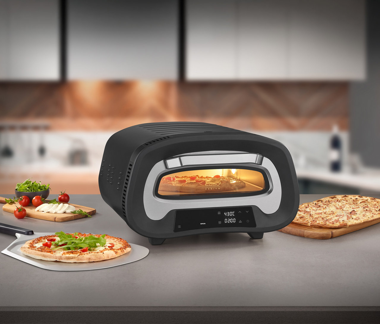 Der PizzaChef 430° Ofen mit Pizza darin steht auf einer Küchentheke. Daneben liegen zwei weitere Pizzen und Zutaten.