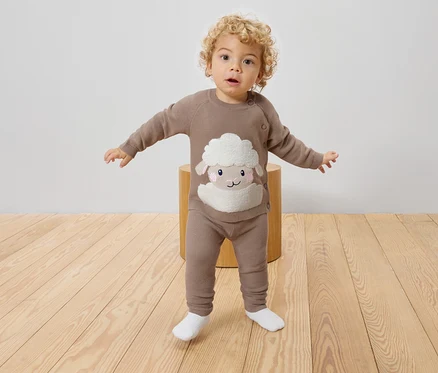 Ein Kleinkind mit lockigem Haar steht auf einem Hocker und trägt einen braunen Strickpullover und Leggings mit einem Schaf darauf.