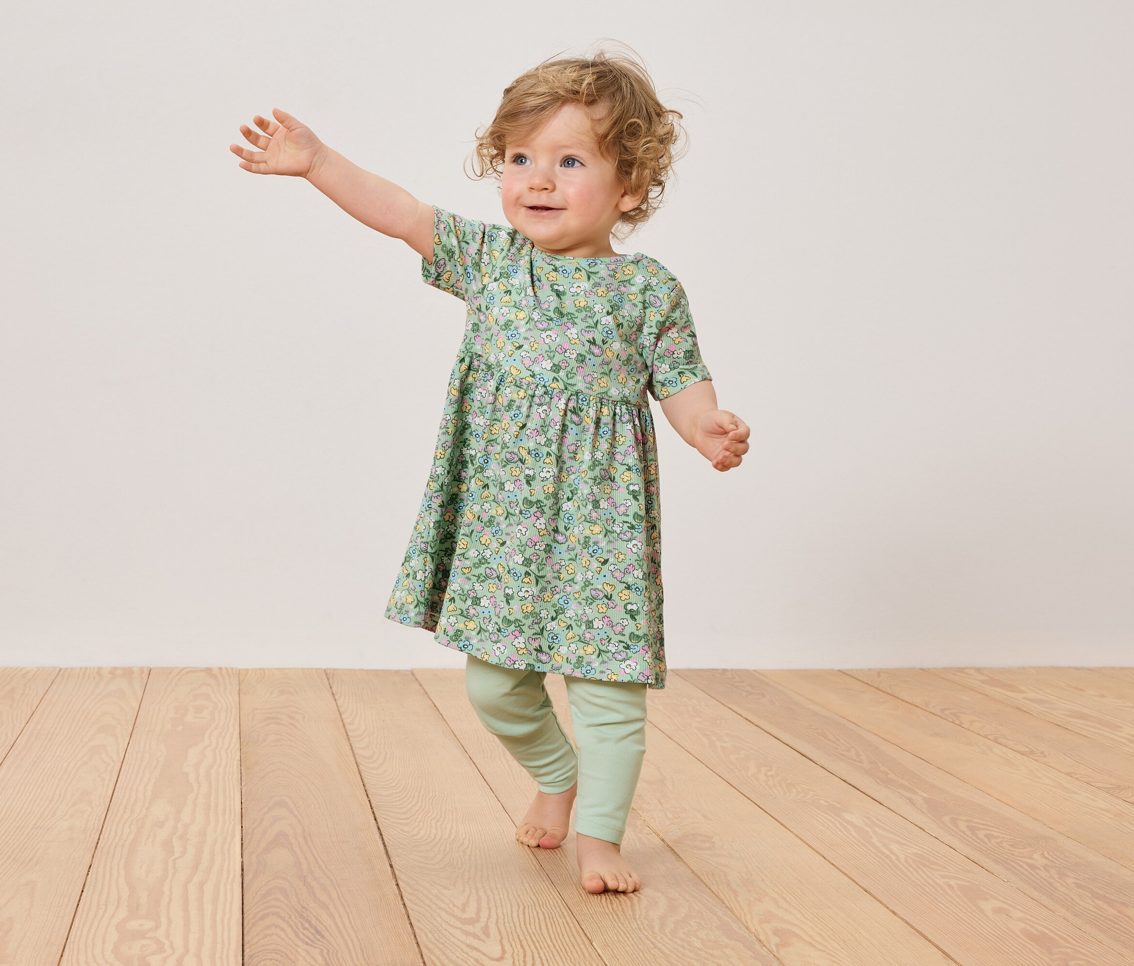 Ein Kleinkind mit blonden Locken trägt ein geblümtes Kleid und grüne Leggings, während es auf einem Holzboden steht.