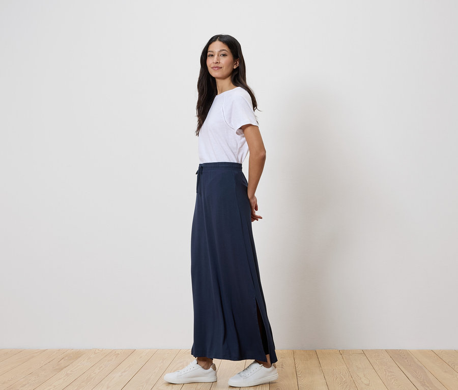 Frau steht seitlich, trägt ein weißes T-Shirt, einen langen marineblauen Rock und weiße Turnschuhe.