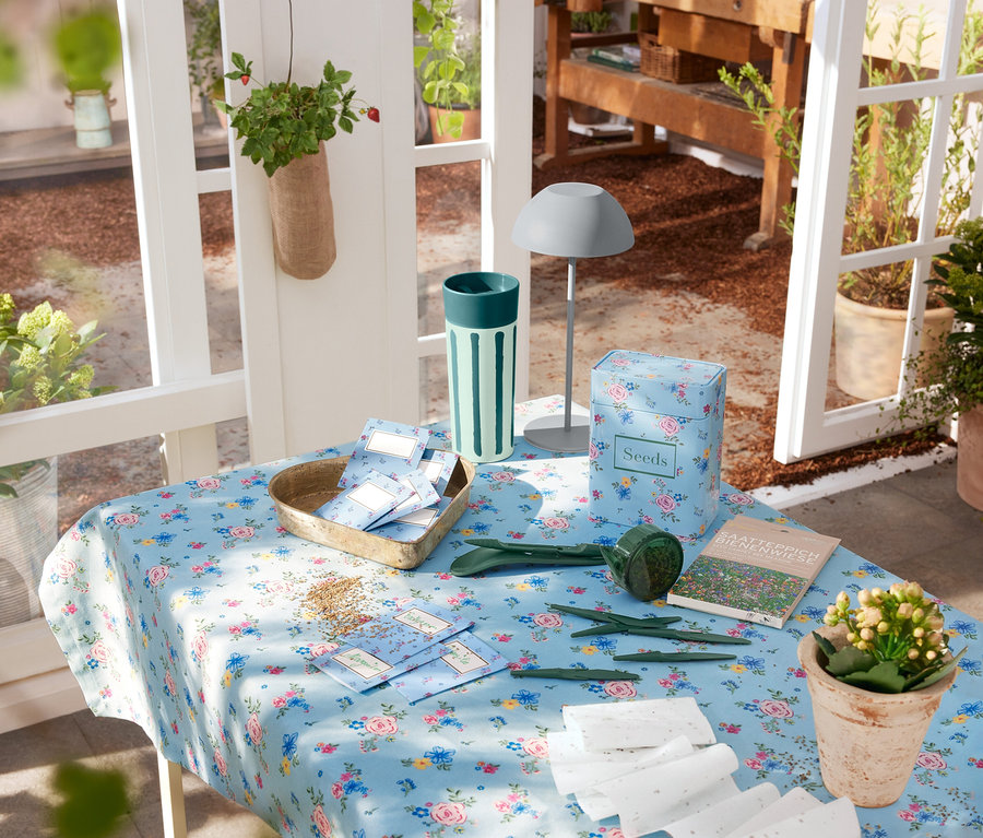 Eine graue LED-Akku-Tischleuchte steht auf einem Tisch mit Blumendecke, neben Gartengeräten und Samen.