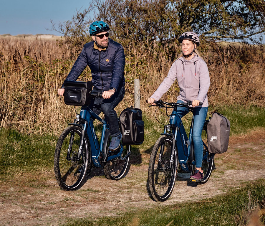 Zwei Personen fahren mit E-Bikes, beide ausgestattet mit FISCHER Gepäckträgertaschen PLUS+ Terra.