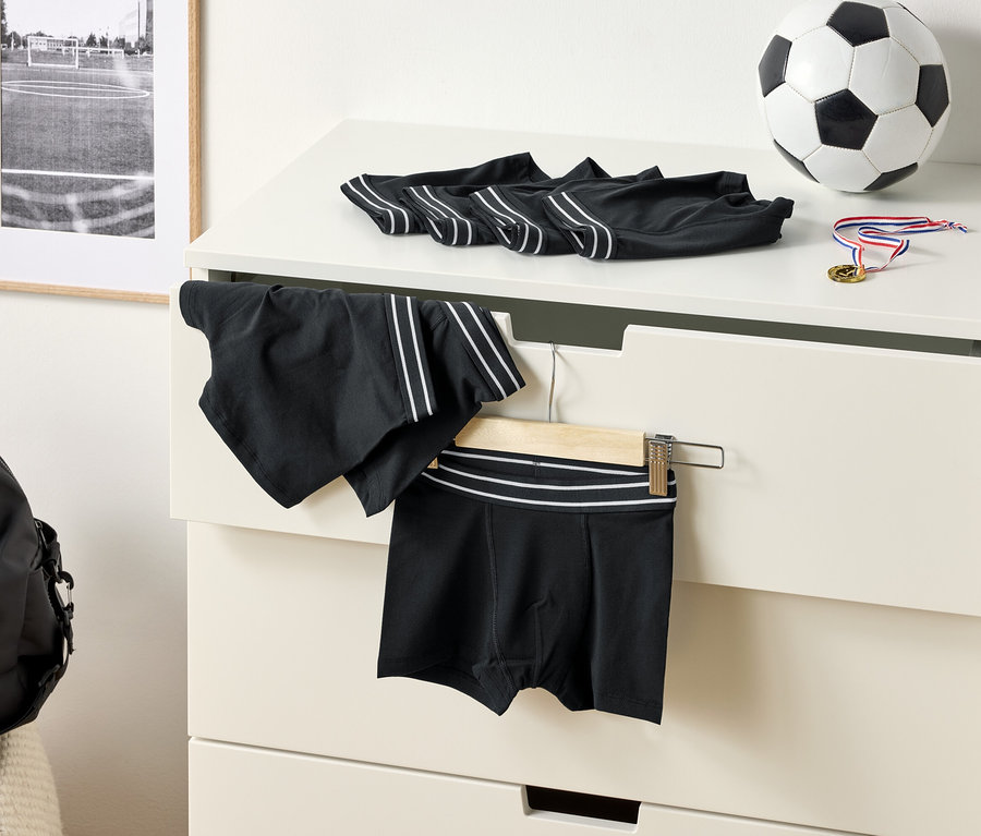 Auf einer Kommode liegen schwarze Unterhosen mit weißen Streifen, ein Fußball und eine Medaille. Eine Sporthose hängt an einem Kleiderbügel.