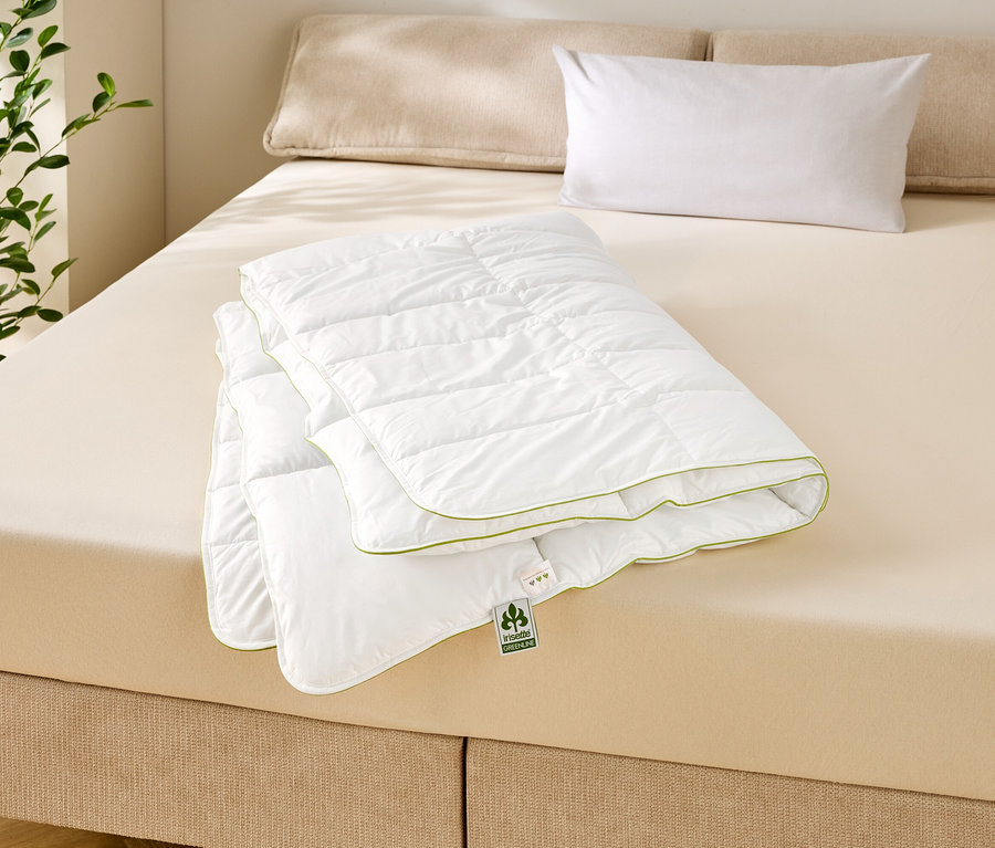 Gefaltete, gesteppte, weiße Decke mit grüner Umrandung auf einem beigen Bett mit zwei Kissen und einer Pflanze im Hintergrund.