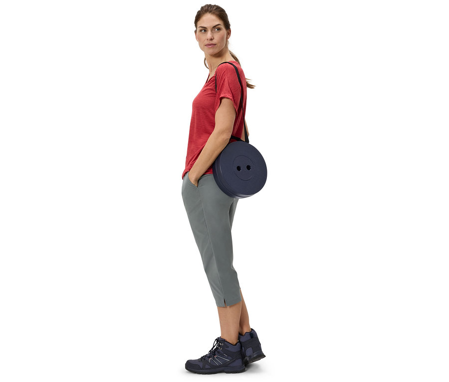 Frau mit rotem T-Shirt und grauen Hosen, die einen runden, marineblauen Hocker über der Schulter trägt.