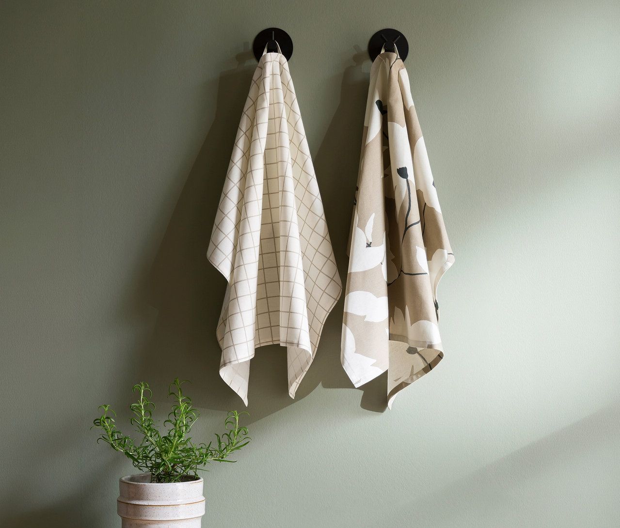 Zwei Geschirrtücher hängen an schwarzen Haken an einer grünen Wand. Das linke Tuch ist cremefarben mit einem hellen Karomuster, das rechte Tuch ist beige mit einem weiß-schwarzen Blumenmuster. Darunter steht eine Rosmarinpflanze im Topf.