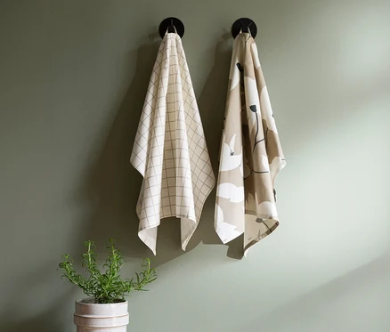 Zwei Geschirrtücher hängen an schwarzen Haken an einer grünen Wand. Das linke Tuch ist cremefarben mit einem hellen Karomuster, das rechte Tuch ist beige mit einem weiß-schwarzen Blumenmuster. Darunter steht eine Rosmarinpflanze im Topf.