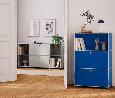 Eine offene weiße Tür verbindet zwei Räume mit Holzböden. Im Vordergrund steht ein blaues modulares Aufbewahrungsmöbel mit einem offenen oberen Regal, das Bücher, einen Diffusor, eine Lampe und eine Pflanze enthält, sowie zwei geschlossenen unteren Fächern. Im Hintergrund steht ein hellgraues modulares Aufbewahrungsmöbel mit offenen und geschlossenen Fächern, das Bücher und eine Pflanze enthält, mit zwei gerahmten abstrakten Kunstwerken darüber.