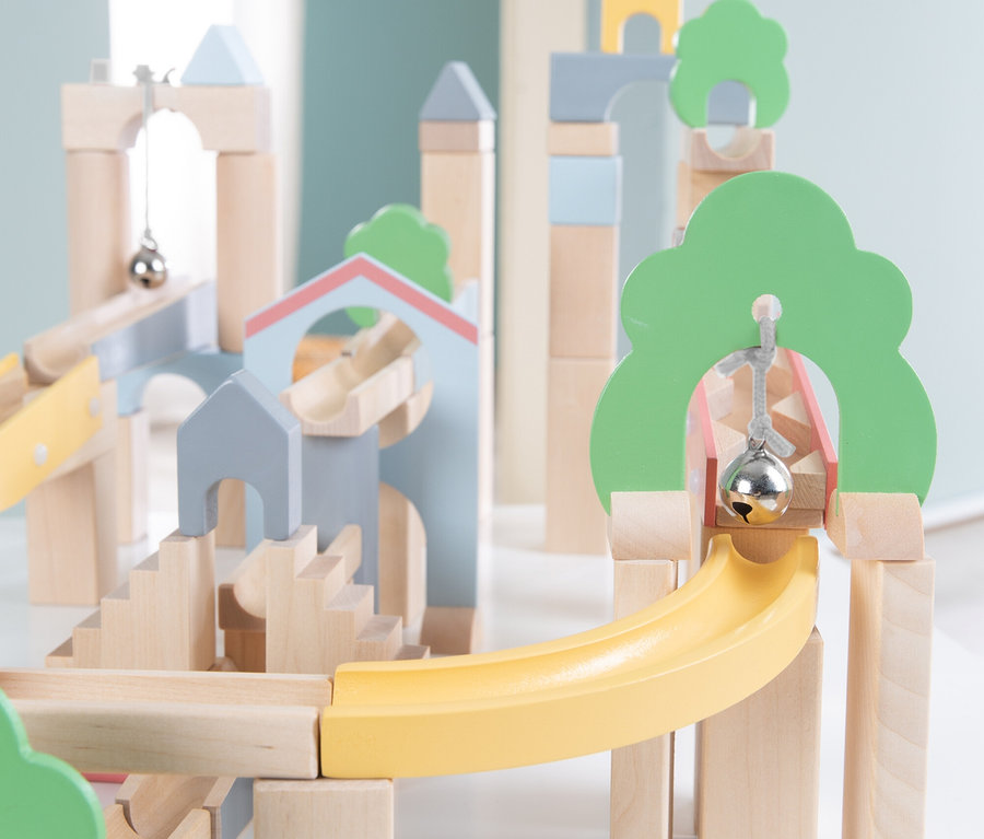 Eine mehrstöckige Murmelbahn aus Holz, gebaut mit Naturholzklötzen, hellgrünen Baumformen, hellblauen und grauen hausähnlichen Strukturen und gelben Rampen. Silberne Glocken hängen an Schnüren in einigen Bögen, und treppenartige Elemente sind sichtbar.