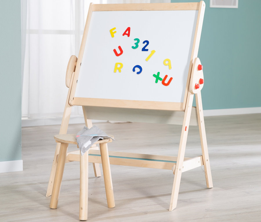 Holzstaffelei mit weißer Tafel, auf der bunte Magnetbuchstaben und -zahlen haften, daneben ein kleiner Holzhocker mit grauem Tuch auf hellem Boden.