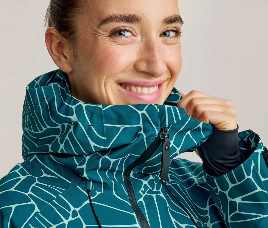 Lächelnde Frau in einer petrolfarbenen Jacke mit hellblauem, abstraktem Muster, die den Kragen hochzieht.