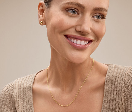 Porträt einer lächelnden blonden Frau, die ein beiges Oberteil, eine goldene Halskette und Ohrringe trägt.