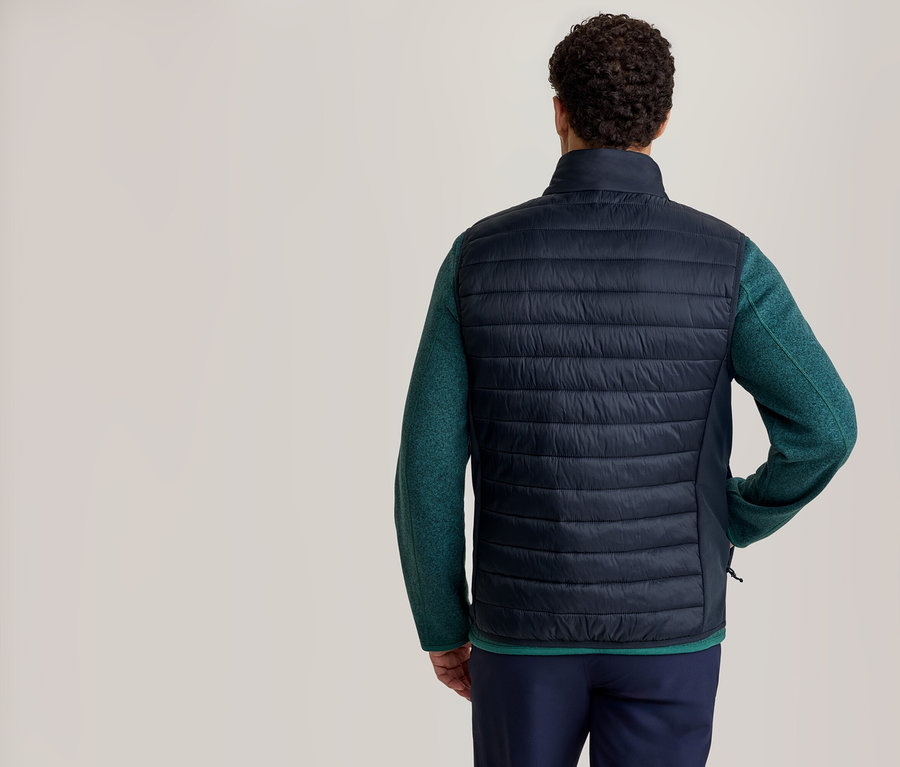Ein Mann von hinten, der eine dunkelblaue Steppweste über einem grünen Langarmshirt und einer dunkelblauen Hose trägt.