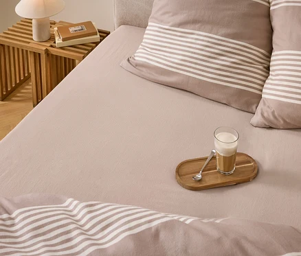 Ein Bett mit hellbrauner Bettwäsche, gestreiften Kissen und Decke. Ein Glas Latte Macchiato mit Löffel auf einem Holztablett liegt auf dem Bett. Daneben ein Nachttisch mit Lampe und Büchern.