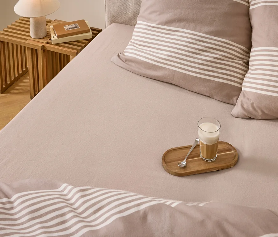 Ein Bett mit hellbrauner Bettwäsche, gestreiften Kissen und Decke. Ein Glas Latte Macchiato mit Löffel auf einem Holztablett liegt auf dem Bett. Daneben ein Nachttisch mit Lampe und Büchern.