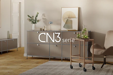 Modernes Wohnzimmer mit großem, taupefarbenem modularem Sideboard, dekoriert mit Vase, Lampe und Kunst. Rechts ein Servierwagen mit Wasserkaraffe und Sessel auf einem Teppich. Links ein kleineres Sideboard. Text: CN3 serie.