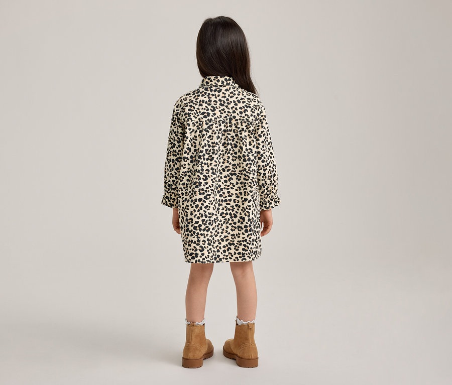 Ein Kind von hinten gesehen, trägt ein langärmeliges Hemdblusenkleid mit Leopardenmuster in Schwarz-Weiß. Das Kind trägt auch hellbraune Stiefel und weiße Rüschensocken.