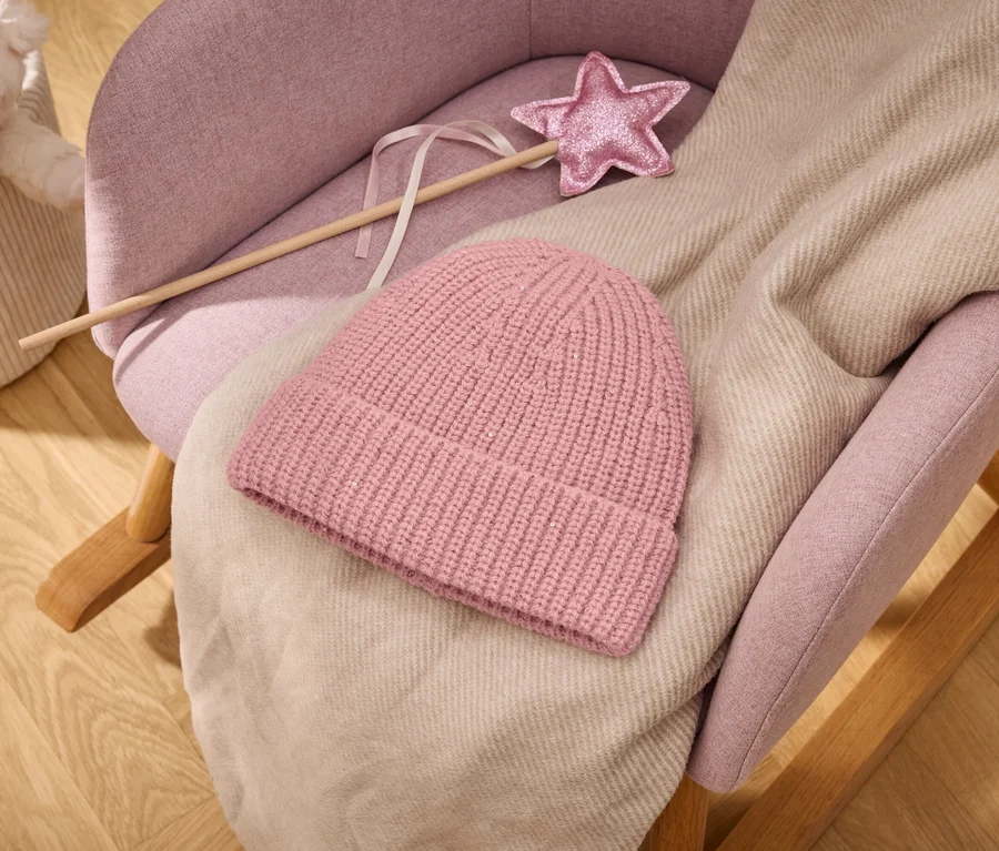 Rosa Strickmütze und hellbeige Decke auf einem rosa gepolsterten Schaukelstuhl mit einem rosa Sternenzauberstab; im Hintergrund ein Korb mit Stofftier auf Holzboden.