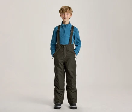 Lächelnder Junge in einem blauen Fleece-Pullover und dunkelgrünen Skihosen mit Hosenträgern, die Hände in den Taschen.