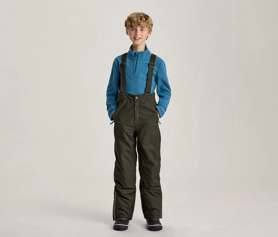 Lächelnder Junge in einem blauen Fleece-Pullover und dunkelgrünen Skihosen mit Hosenträgern, die Hände in den Taschen.