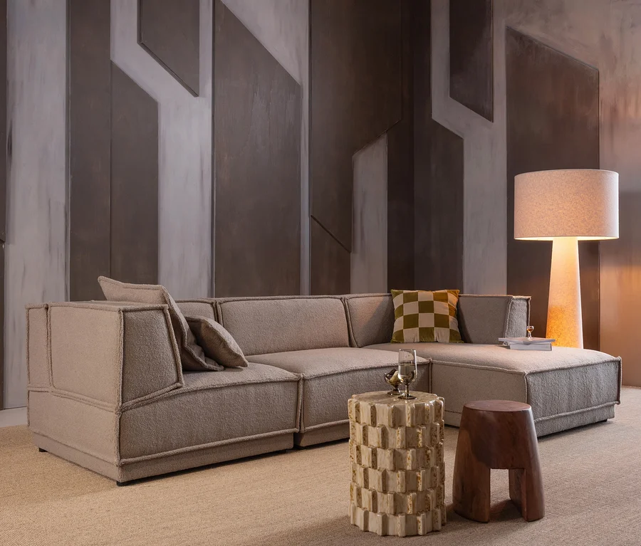 Beiges L-förmiges Sofa mit Kissen, darunter ein grün kariertes, in einem modernen Wohnzimmer. Ein zylindrischer Beistelltisch, ein Holzhocker und eine Stehlampe stehen auf hellem Teppich vor einer geometrischen Wand.