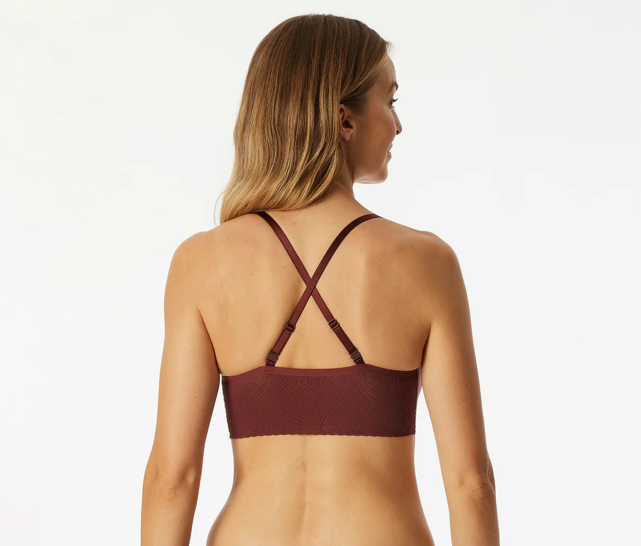Frau von hinten, trägt einen bordeauxroten Bralette mit gekreuzten Trägern. Ihr blondes Haar fällt über ihre Schultern.