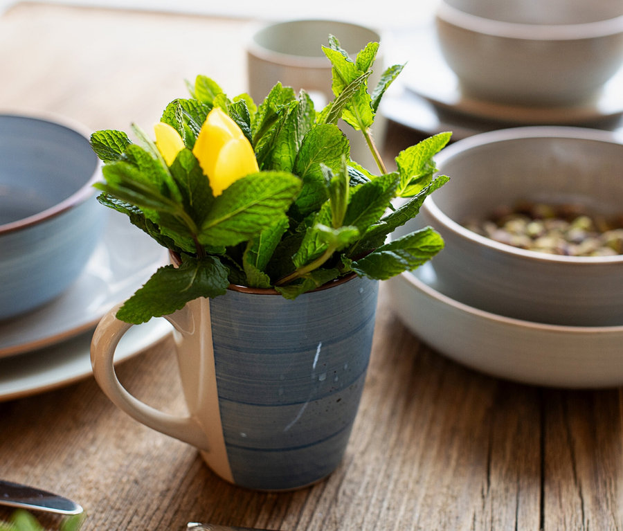 Blau-beige Tasse mit Minze und Zitronenspalte auf Holztisch, umgeben von blauem und grauem Geschirr des Mäser Geschirr-Sets »Valmera«.
