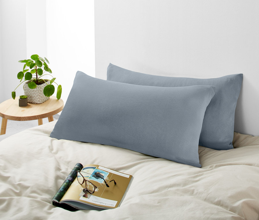 Zwei hellgraue Jersey- Kopfkissenbezüge liegen auf einem Bett neben einer Zeitschrift und einer Brille.