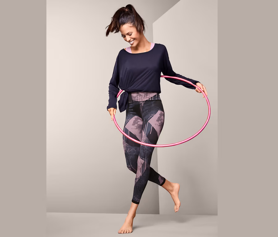 Frau lächelt und dreht einen pinken Hula-Hoop-Reifen um ihre Taille, sie trägt eine Wende-Sporttight.