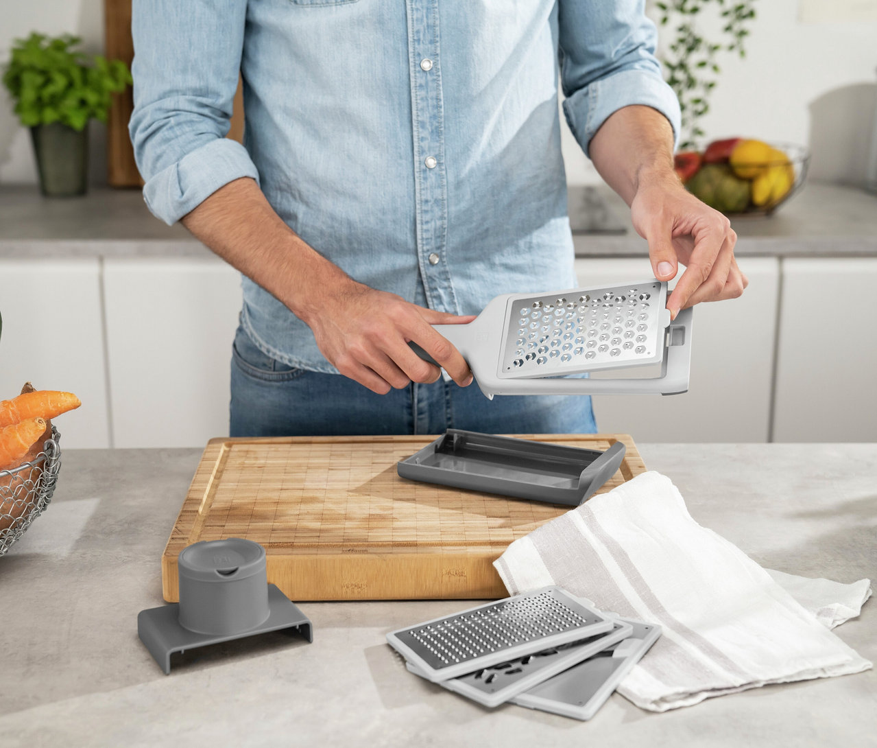 Eine Person in Jeanshemd hält eine graue ZWILLING Z-Cut Multireibe und setzt einen Reibeeinsatz ein. Auf der Arbeitsplatte liegen weitere Reibeeinsätze und ein Holzschneidebrett.