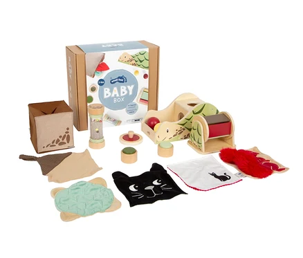 Eine Sammlung von Babyspielzeug, darunter eine "BABY BOX" aus Karton, ein Holzspielzeug in Schildkrötenform, eine Rassel, Stapelspielzeug und verschiedene Stoffartikel wie eine schwarze Katze und eine rote Felltextur.