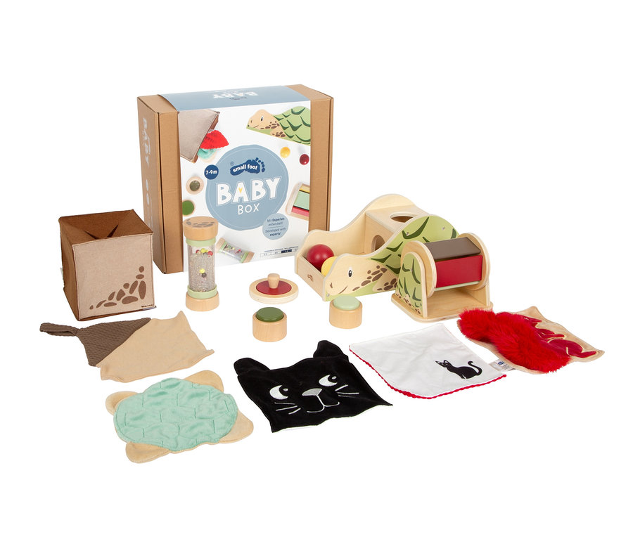 Eine Sammlung von Babyspielzeug, darunter eine "BABY BOX" aus Karton, ein Holzspielzeug in Schildkrötenform, eine Rassel, Stapelspielzeug und verschiedene Stoffartikel wie eine schwarze Katze und eine rote Felltextur.