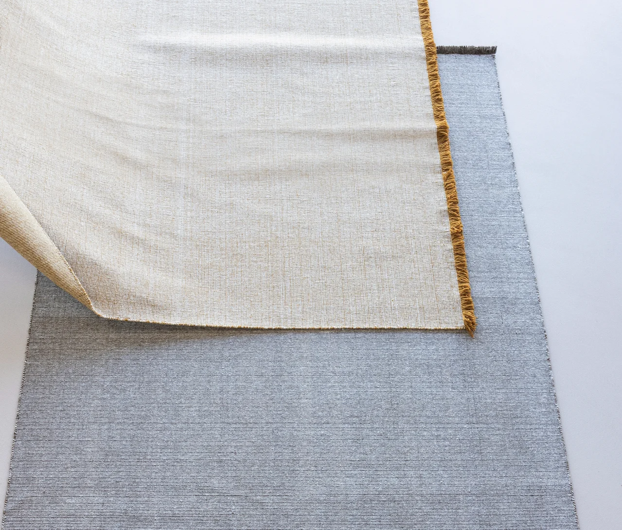 Ein hellbrauner WOOOD Teppich »Weavly« ist teilweise über einem grauen Teppich aufgerollt und zeigt seine gewebte Textur und Fransen.