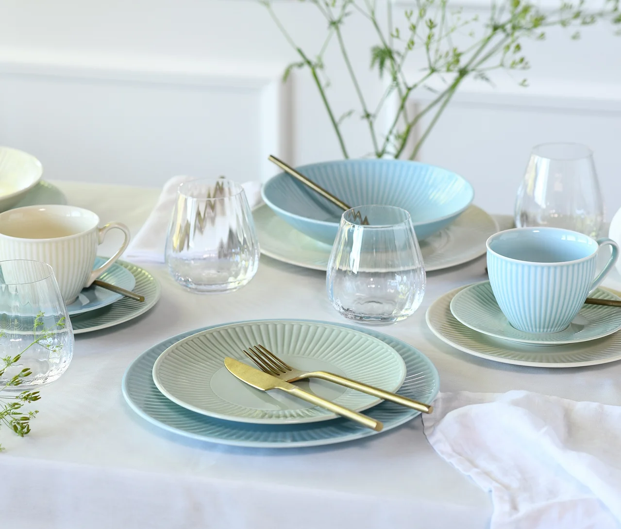 Ein gedeckter Esstisch mit pastellfarbenem creaTable Kombiservice »Valencia Pastell«. Der Tisch ist mit einem weißen Tischtuch bedeckt und zeigt hellblaue, hellgrüne und cremefarbene gerippte Teller, Schalen und Teetassen mit Untertassen. Goldfarbenes Besteck und klare Gläser sind ebenfalls vorhanden. Im Hintergrund steht eine Vase mit grünem Laub.