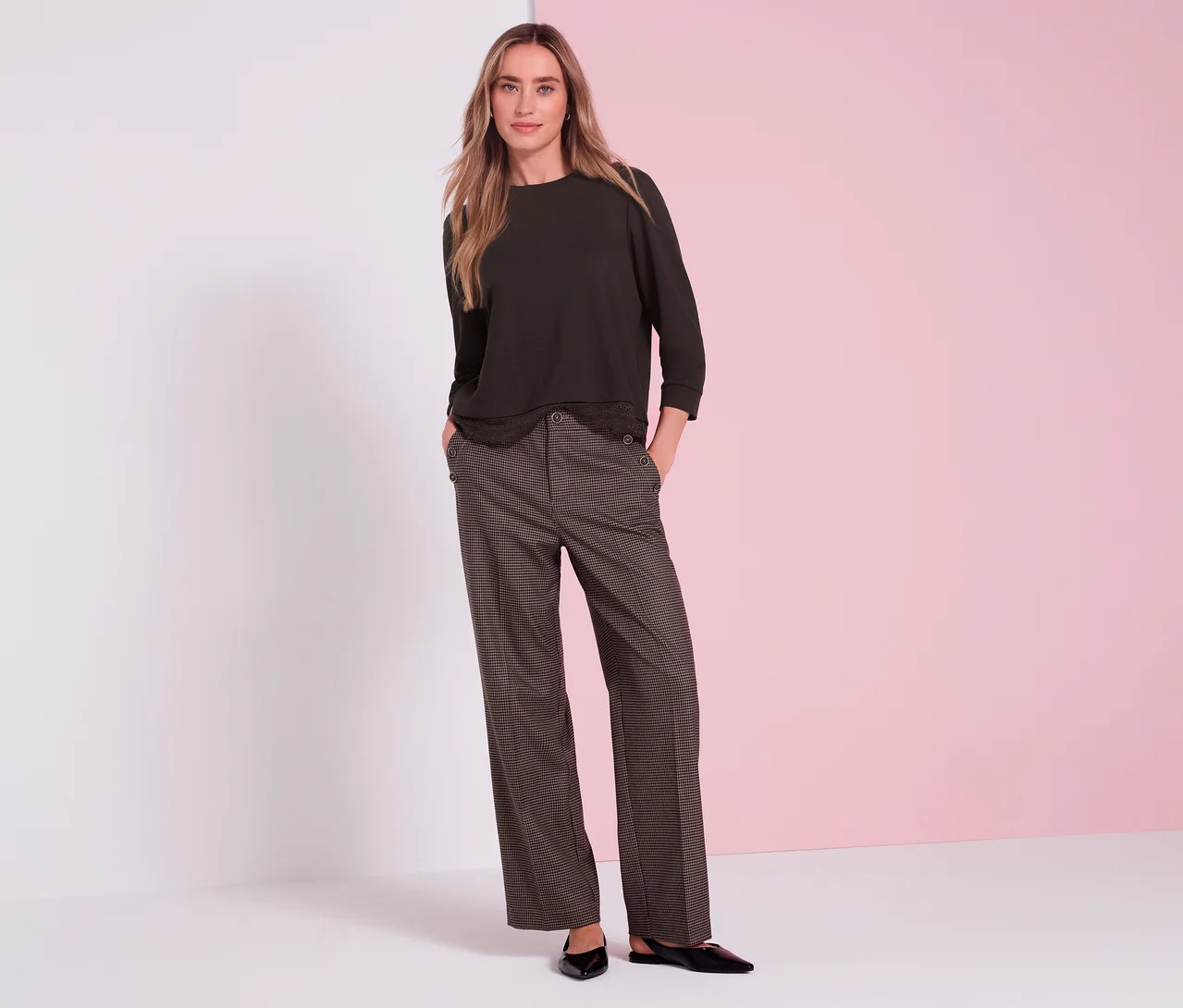 Eine Frau mit langen blonden Haaren steht mit den Händen in den Taschen und trägt ein braunes Street One 3/4-Jerseyshirt, braun gemusterte weite Hosen mit Zierknöpfen an den Taschen und schwarze flache Schuhe vor einem weiß-rosa Hintergrund.