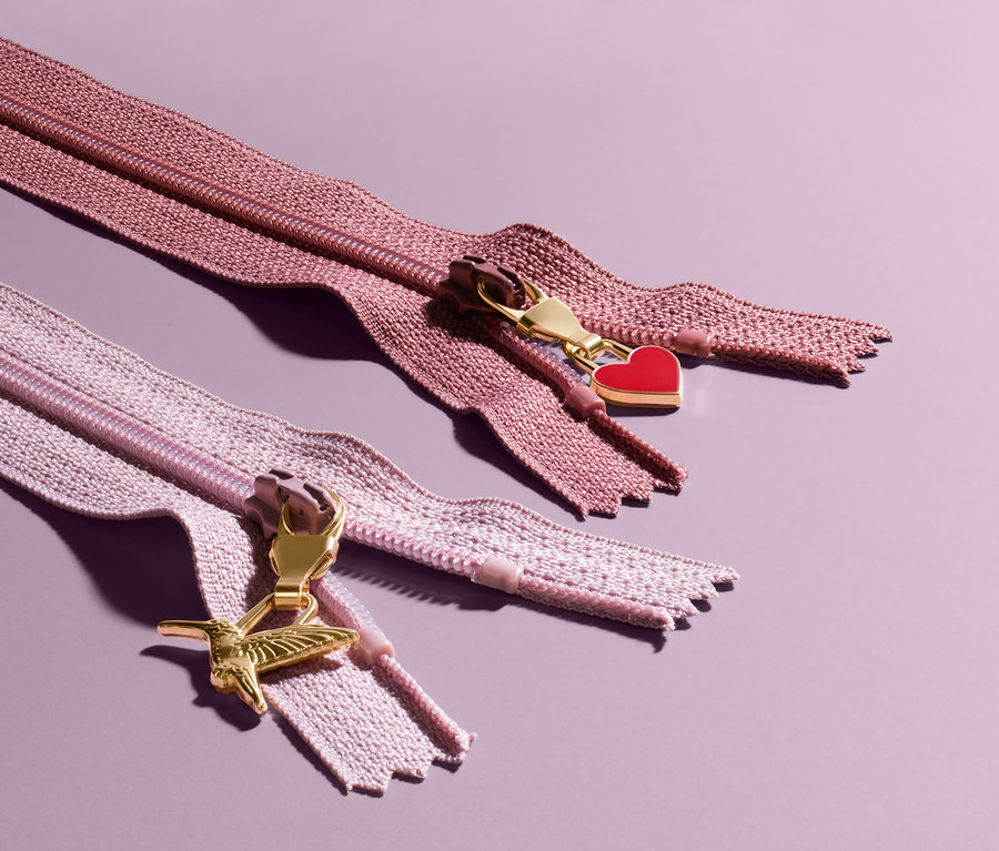 Zwei Reißverschlüsse mit dekorativen Anhängern auf violettem Hintergrund. Der obere Reißverschluss hat einen roten herzförmigen Anhänger, der untere einen kolibri-förmigen. 6 Fashion-Reißverschlussanhänger.