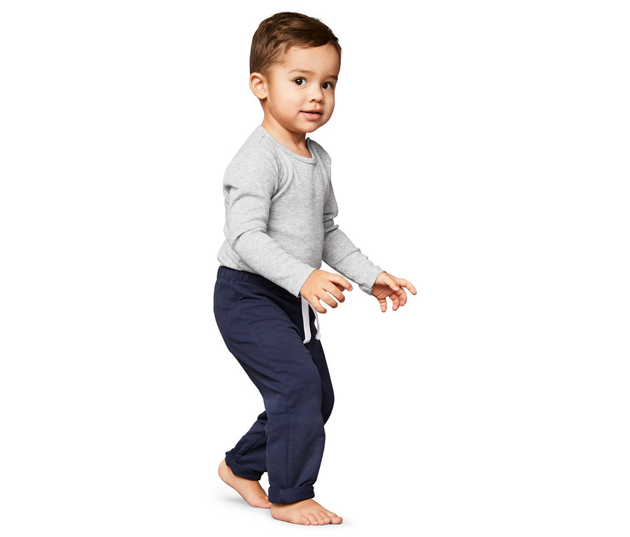 Ein Junge in grauem Oberteil und marineblauen 2 Schlupfhosen steht barfuß vor einem weißen Hintergrund.