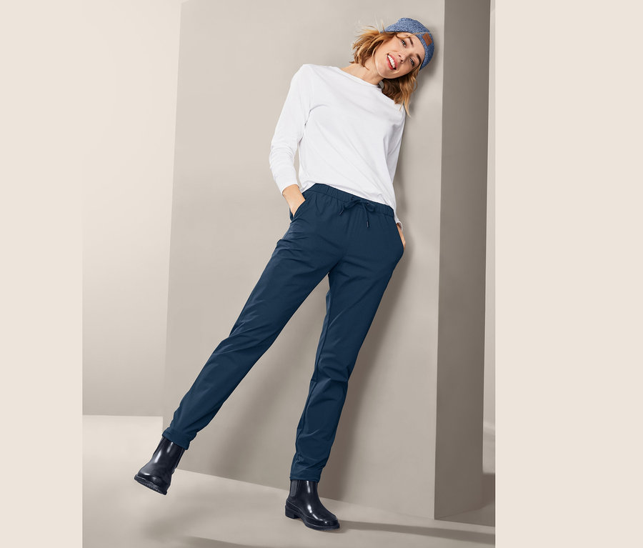 Frau posiert in Softshell-Joggpants, einem weißen Oberteil und schwarzen Schuhen.