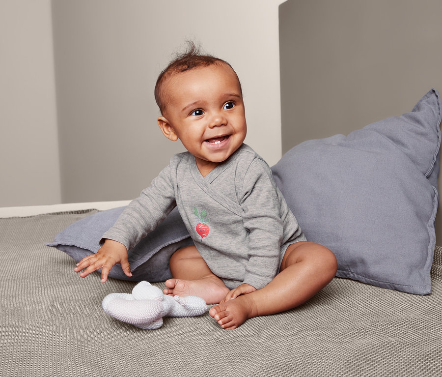 Lächelndes Baby sitzt auf einer grauen Decke und trägt einen grauen Langarmbody mit Radieschen-Print.