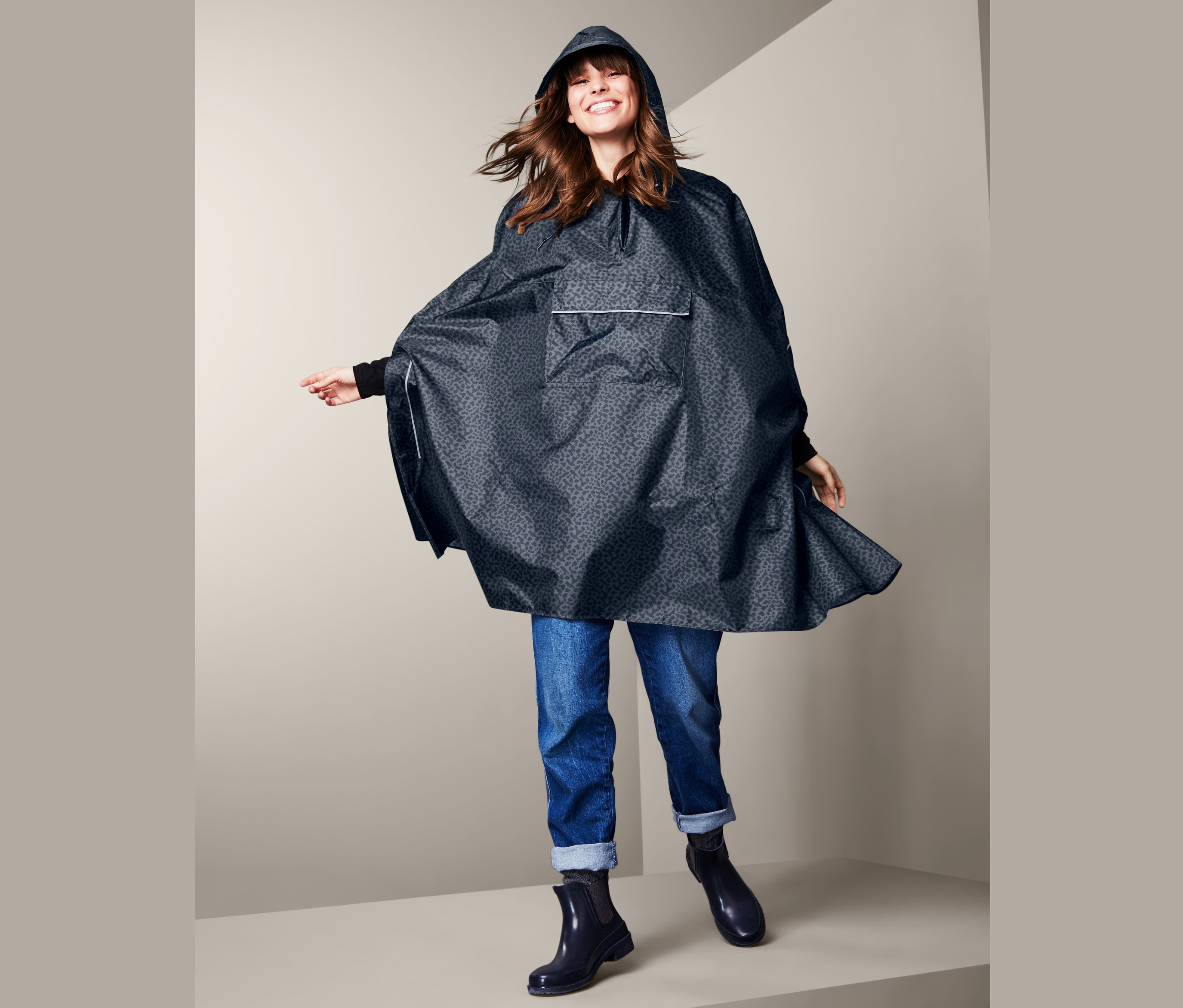 Lächelnde Frau posiert in blauem Regenponcho mit Kapuze, Jeans und Stiefeln.