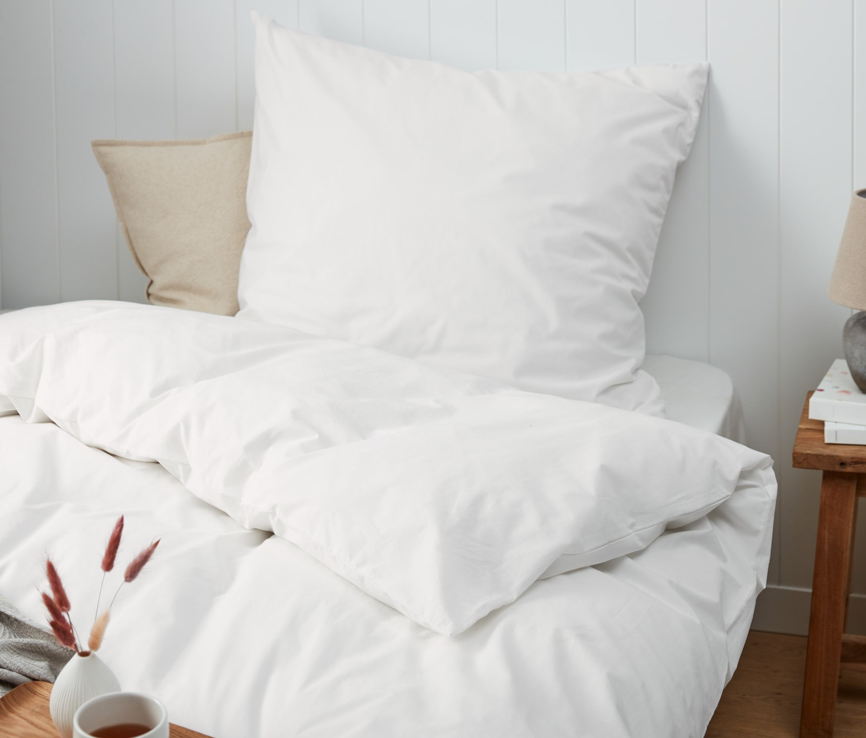 Weisse Perkal-Bettwäsche in Normal- und Übergrösse auf einem Bett mit einem Holztablett, einer Tasse und einer Vase.