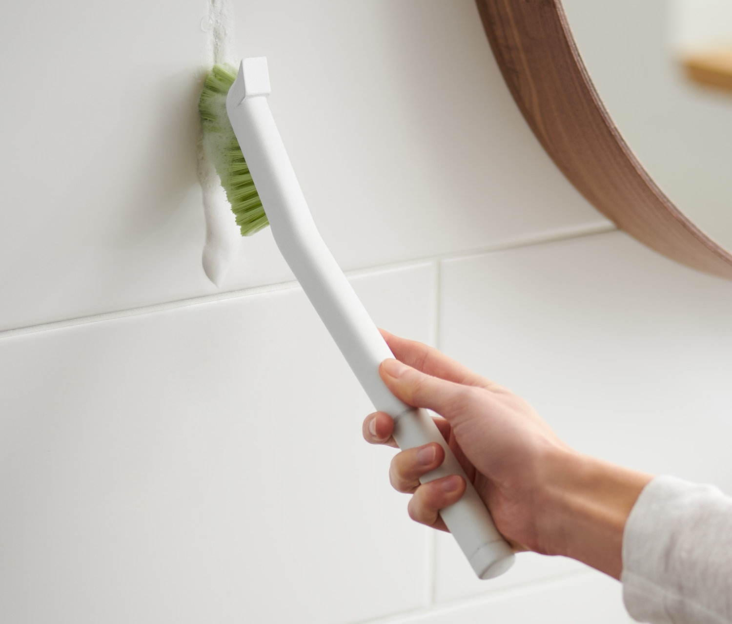 Eine Frau reinigt die Fugen zwischen weißen Fliesen mit einer langen Fugenbürste.