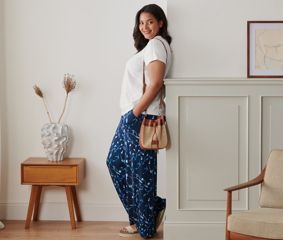 Eine Frau lehnt an einer Wand und posiert in einer Palazzohose, einem weißen Shirt und Plateau-Sandale aus Leder. Sie hat eine Beuteltasche über der Schulter.