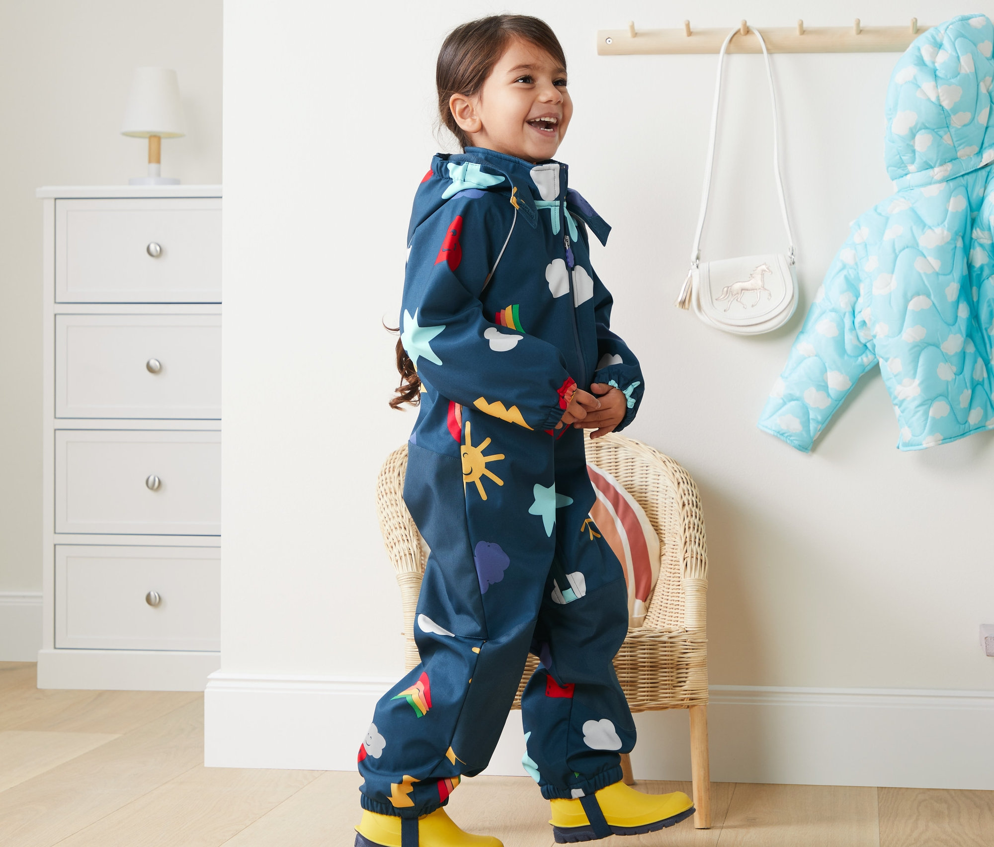 Ein lächelndes Mädchen geht in einem Softshell-Einteiler aus recyceltem Material mit Regenbogen-, Wolken- und Sternmotiven.