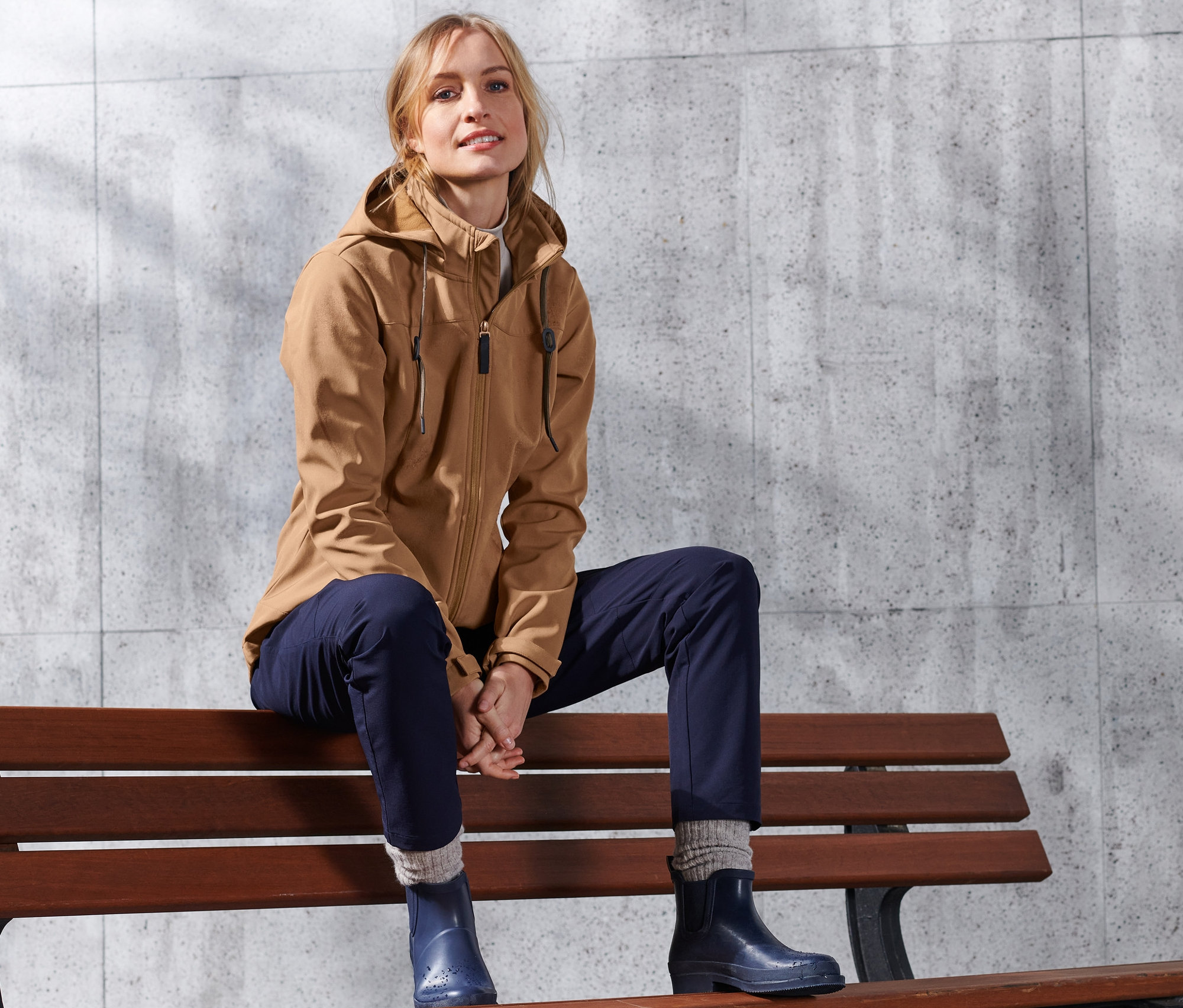 Blonde Frau sitzt auf einer Bank und trägt eine cognacfarbene Softshell-Jacke.